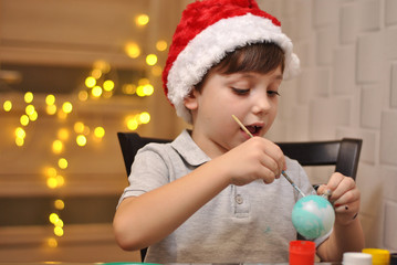 Fototapeta premium Little cute kid decorating Christmas balls at home kitchen on the yellow lights background