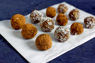 Raw food balls.Handmade vegan sweets made from almonds, walnuts, dates, raisins, coconut and cocoa. Raw food candies in coconut and cocoa close-up on white paper on a dark table laid out in rows. 