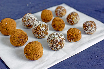 Raw food balls.Handmade vegan sweets made from almonds, walnuts, dates, raisins, coconut and cocoa. Raw food candies in coconut and cocoa close-up on white paper on a dark table laid out in rows. 