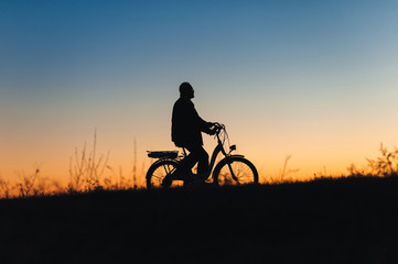 Fototapeta na wymiar Male cyclist on the e-bike or electric bicycle on the sunset background riding up the hill. Silhouette of the old man in profile. Active pension. Travel. Sport.