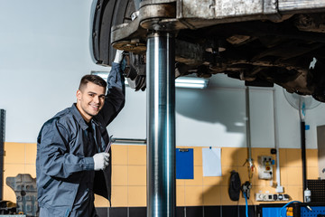 Fototapeta na wymiar smiling mechanic holding wrench and looking at camera while standing under car raised on car lift