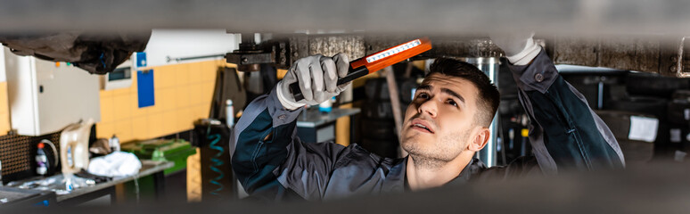 Fototapeta premium panoramic shot of mechanic inspecting car bottom with flashlight