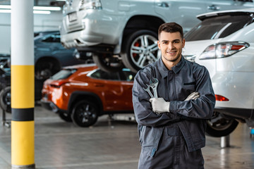smiling mechanic looking at camera while standing with crossed arms and holding wrenches