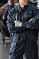 cropped view of young mechanic standing with crossed arms and holding wrenches