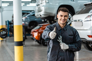 cheerful mechanic looking at camera and showing thumb up while holding wrenches - obrazy, fototapety, plakaty