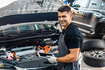 handsome mechanic smiling at camera while using digital tablet near car engine compartment