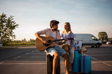 Handsome bearded young man playing guitar music for a beautiful young blonde. They are waiting for transportation to their tourist destination. Transport. Travel.