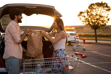 A young couple is in the parking lot of a large shopping mall, they just finished buying food and...
