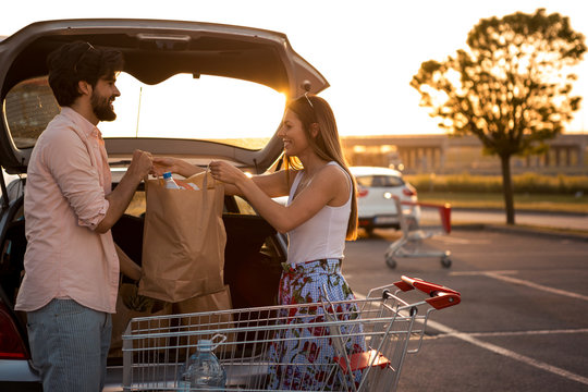 A young couple is in the parking lot of a large shopping mall, they just finished buying food and loading their food into the trunk of a car. Food. Shopping.