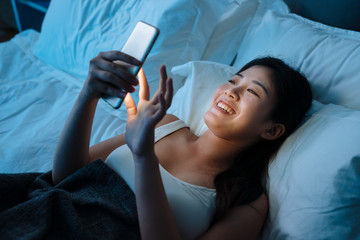 young beautiful asian japanese woman in bed using mobile phone late at night dark bedroom. happy...