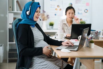 Cute pregnant muslim woman talking by headset in office. maternity businesswoman in suit having conversation online solving customer problem in workplace. asian chinese female colleague in back