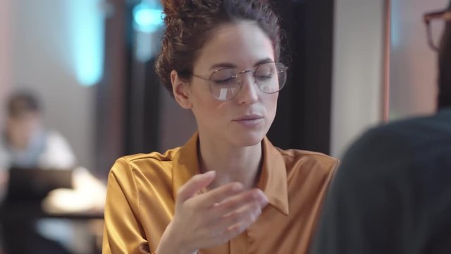 Over-shoulder chest-up shot of young Caucasian woman with curly hair, in glasses sitting at table in restaurant with black boyfriend and talking to him about difficult day at work
