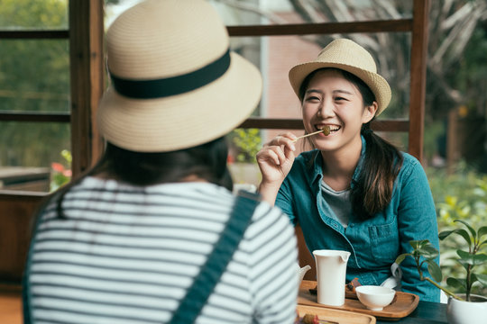 Smiling Asian Korean Female Friends Drinking Matcha Tea And Laughing. Lady In Straw Hat Eating Plum Snack And Laughing Listening To Sister Talking. Japanese Architecture Style House Cafe Kyoto Japan