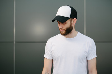 Hipster handsome male model with beard wearing white blank t-shirt and a baseball cap with space for your logo or design in casual urban style