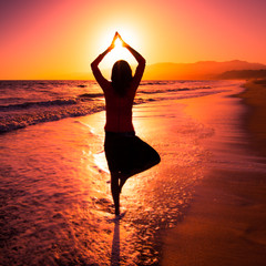 Girl silhouette in yoga Vrksasana Tree Pose balancing on one leg in ocean catches last sunsetting rays in hands above her head. Sun greetings, meditation in nature, healthy summer relax exercise ideas
