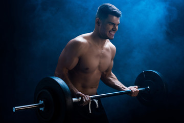 sexy muscular bodybuilder with bare torso excising with barbell on black background with smoke