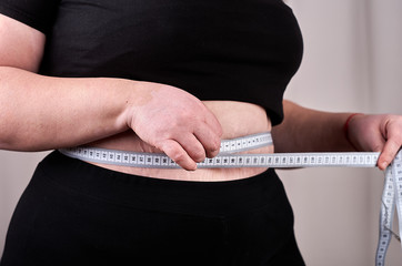 Chubby woman standing and using measuring tape for her waist in a room.