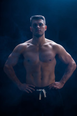 Fototapeta na wymiar sexy muscular bodybuilder with bare torso posing with hands on hips on black background with smoke