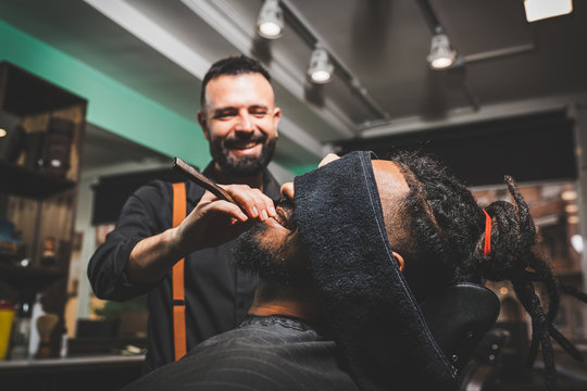barbero blanco caucasico arreglando, cortando y afeitando con navaja de afeitar la barba a un chico afroamericano. barberia y personas de estilo hipster