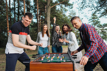 Friends play table football or kicker outdoors. Players and fans rejoice in the victory. - obrazy, fototapety, plakaty