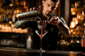 Male bartender pouring a alcoholic drink from the steel shaker to the glass through the sieve