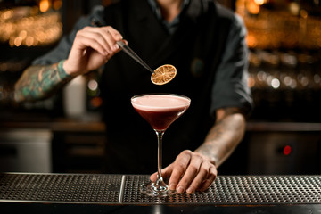 Professional male bartender decorating a delicious alcoholic cocktail in the glass with a dried orange with tweezers