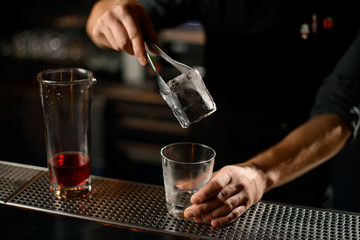 Fototapeta na wymiar Male bartender putting an ice cube with tweezers to the empty cocktail glass