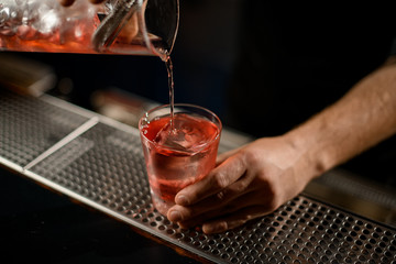 Bartender pouring a alcoholic drink from the measuring cup to the glass through the strainer filter