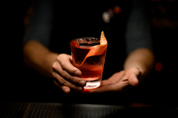 Male bartender serving the red alcoholic drink decorated with the spiral orange zest and ice