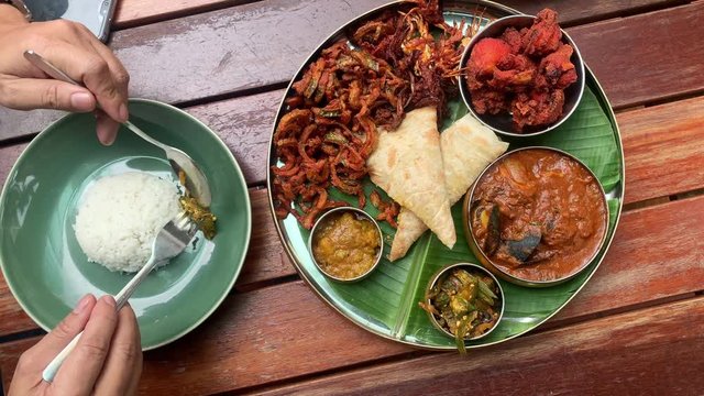 Meals served on banana leaf, consist of fried chicken, curry seafood, canai and etc, a traditional South Indian cuisine. Man’s hand picking up food and put to his plate of rice. Top view.
