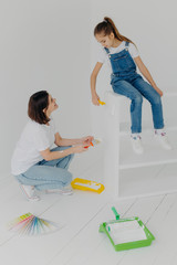 Shot of little girl sits on white furniture, refurbish drawer together with mother, use paint rollers and brush, pose in apartment against white background. Children, parents, repairing concept