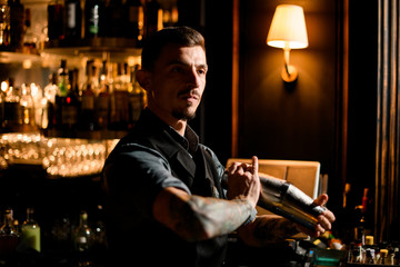 Male bartender with a beard holding in hands a steel shaker ready to shake it in the bar