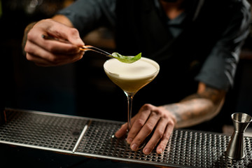 Bartender decorated the yellow creamy color alcoholic cocktail drink with a green leaf with tweezers