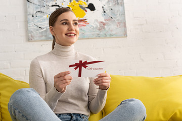 Smiling young woman holding gift voucher and looking away on sofa