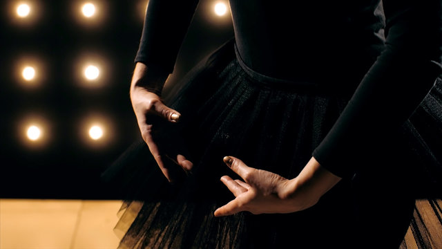 Close-up silhouette of ballerina in black dress is standing in ballet dancing pose. Hands close-up in dark studio with the lights on the background.