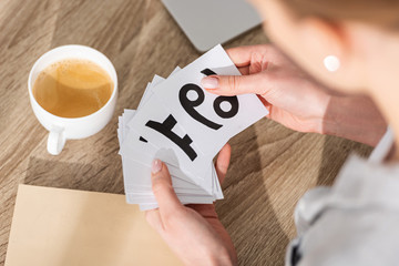 Cropped view of woman holding cards with zodiac signs beside coffee and laptop on table