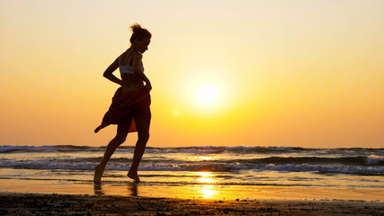 Silhouette of young girl dancing at sunset. Female artist is dancing on the sandy beach against orange sunset.