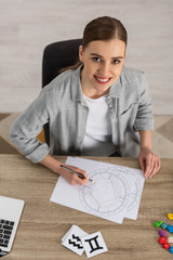 Beautiful smiling astrologer drawing natal chart by cards and stones with zodiac signs on table