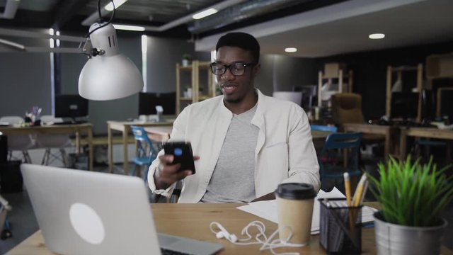 Portrait young african amerian man uses phone and works on laptop at the office