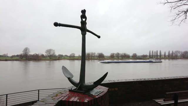 Coastal trading vessel crossing the river on a cloudy day with anchor statue on foreground