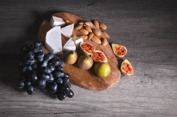 Still life cheese plate on a wooden cutting board lies sliced cheese with mold almonds in the shell and sliced figs next to a bunch of black grapes. Delicious and healthy food concept