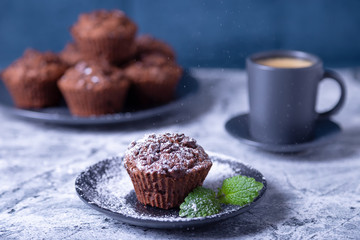 Chocolate muffins with mint on a black plate, strewed with icing sugar. Homemade baking. In the background is a cup of coffee and a plate with muffins. Marble table and blue background. 