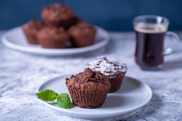 Chocolate muffins with mint on a black plate, strewed with icing sugar. Homemade baking. In the background is a cup of coffee and a plate with muffins. Marble table and blue background. 