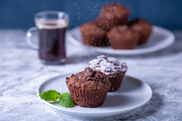 Chocolate muffins with mint on a black plate, strewed with icing sugar. Homemade baking. In the background is a cup of coffee and a plate with muffins. Marble table and blue background. 