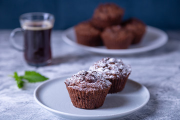 Chocolate muffins with mint on a black plate, strewed with icing sugar. Homemade baking. In the background is a cup of coffee and a plate with muffins. Marble table and blue background. 