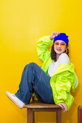 Smiling of happy teen girl 14-19 years in glasses white shirt blue jeans sitting on the wooden chair on yellow background