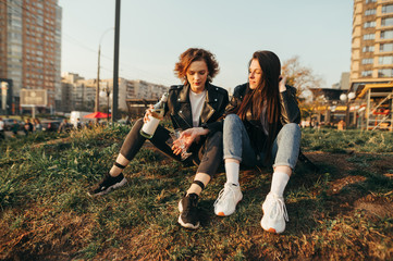 Naklejka na ściany i meble Two cheerful girls in stylish clothes sitting on the grass on a background of cityscape at sunset and drinking wine. Positive girlfriends drink wine on a walk and smile.