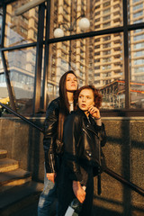 Fototapeta na wymiar Street portrait of two stylish girlfriends in casual clothes, wearing a leather jacket, posing for the camera on an urban background with architecture. Two attractive models in street fashion clothes