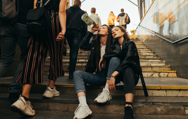 Freedom girls sit on the stairs in a crowd of people and drink wine. Girlfriends drinking alcohol from a bottle sitting on the stairs at the exit of the subway on a background of passersby.