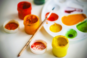 On a white table there are art supplies - a white plastic palette for mixing paints, several colors of gouache paint, a  brush and a lid from a jar of red paint.